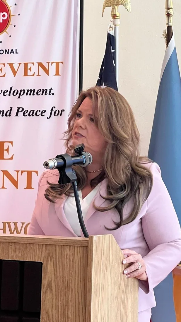 Sherri Combs speaking at a lectern with international flags and a COJEP event banner behind her